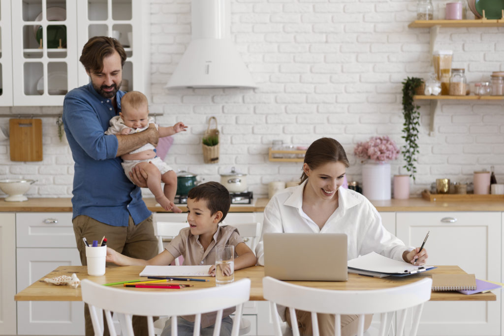 Eine junge Familie hält sich in der Küche auf. Die Frau sitzt am Tisch am Laptop, neben ihr ein älteres Kind. Der Mann steht neben ihr und hält das jüngere Kind auf dem Arm.